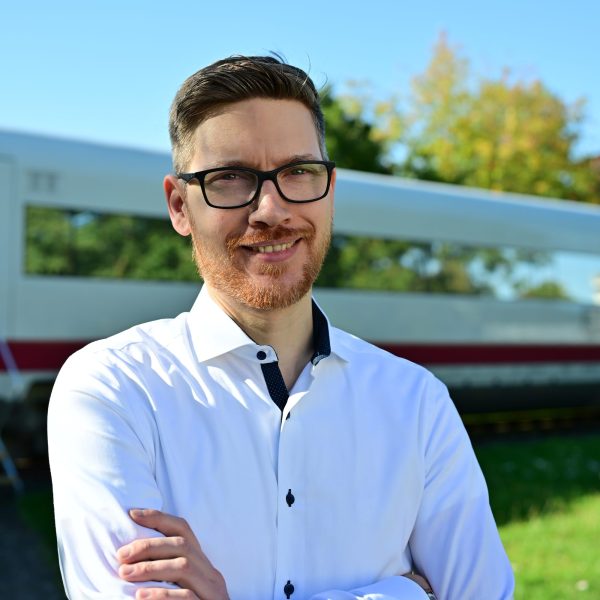 Ein junger Mann der eine Brille trägt, steht bei Sonnenschien vor einem ICE